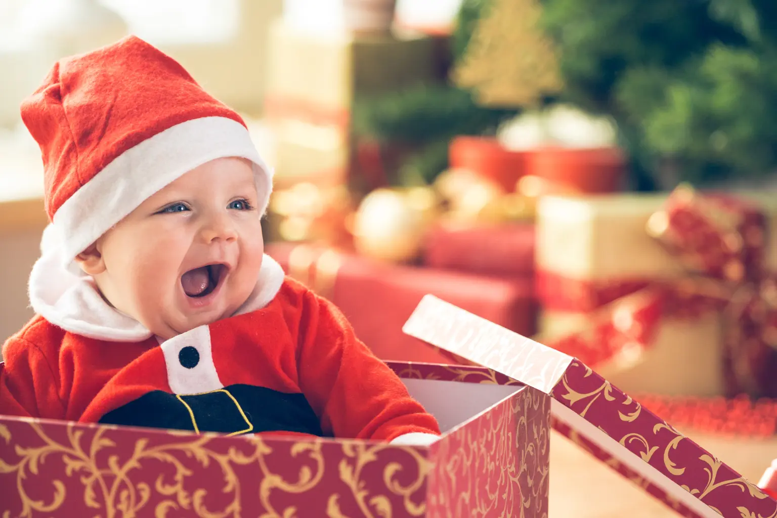 Lachendes Baby mit Weihnachtsmütze sitzt in einem geöffneten Geschenkkarton