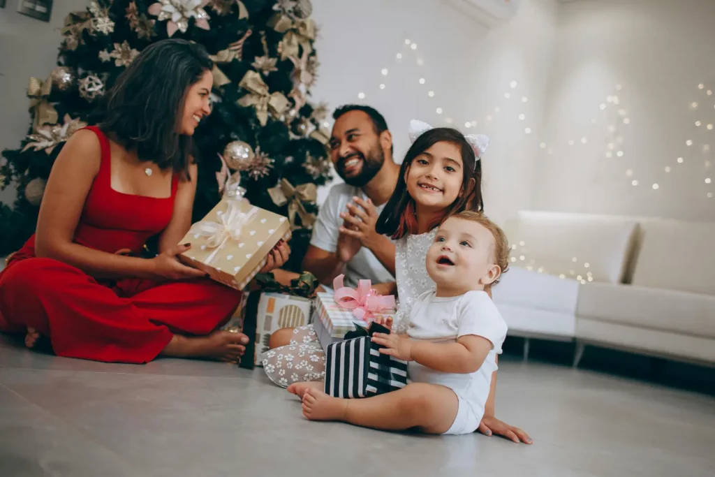 Familie mit Baby und Schwester sitzt lächelnd mit Geschenken vor dem Weihnachtsbaum